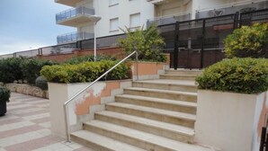 En utomhus trappa som leder upp till en byggnad med ett metallräcke, omgiven av grönska och ett klinkergolv.