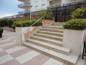 A set of outdoor stairs leading to a building with a metal railing, surrounded by greenery and a tiled floor. - Seafront Apartment with Balcony, Pool, Parking, and WiFi in Torredembarra (Torredembarra)