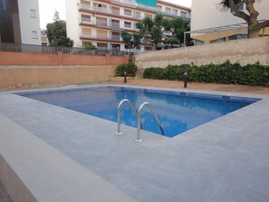 A rectangular swimming pool with two metal ladders, surrounded by a low brick wall, with a multi-story building in the background. - Seafront Apartment with Balcony, Pool, Parking, and WiFi in Torredembarra (Torredembarra)