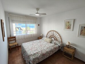 A bedroom with a bed, a chair, a nightstand, a ceiling fan, and a window with a view of the beach. - Seafront Apartment with Balcony, Pool, Parking, and WiFi in Torredembarra (Torredembarra)
