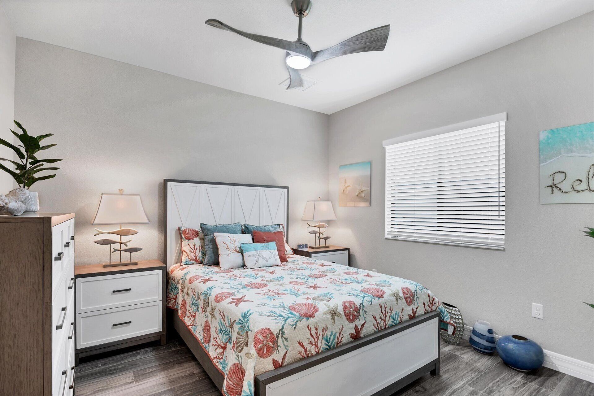 Une chambre à coucher comprenant un lit, des tables de chevet, un ventilateur de plafond, une fenêtre avec des stores et un mur orné d’un tableau.