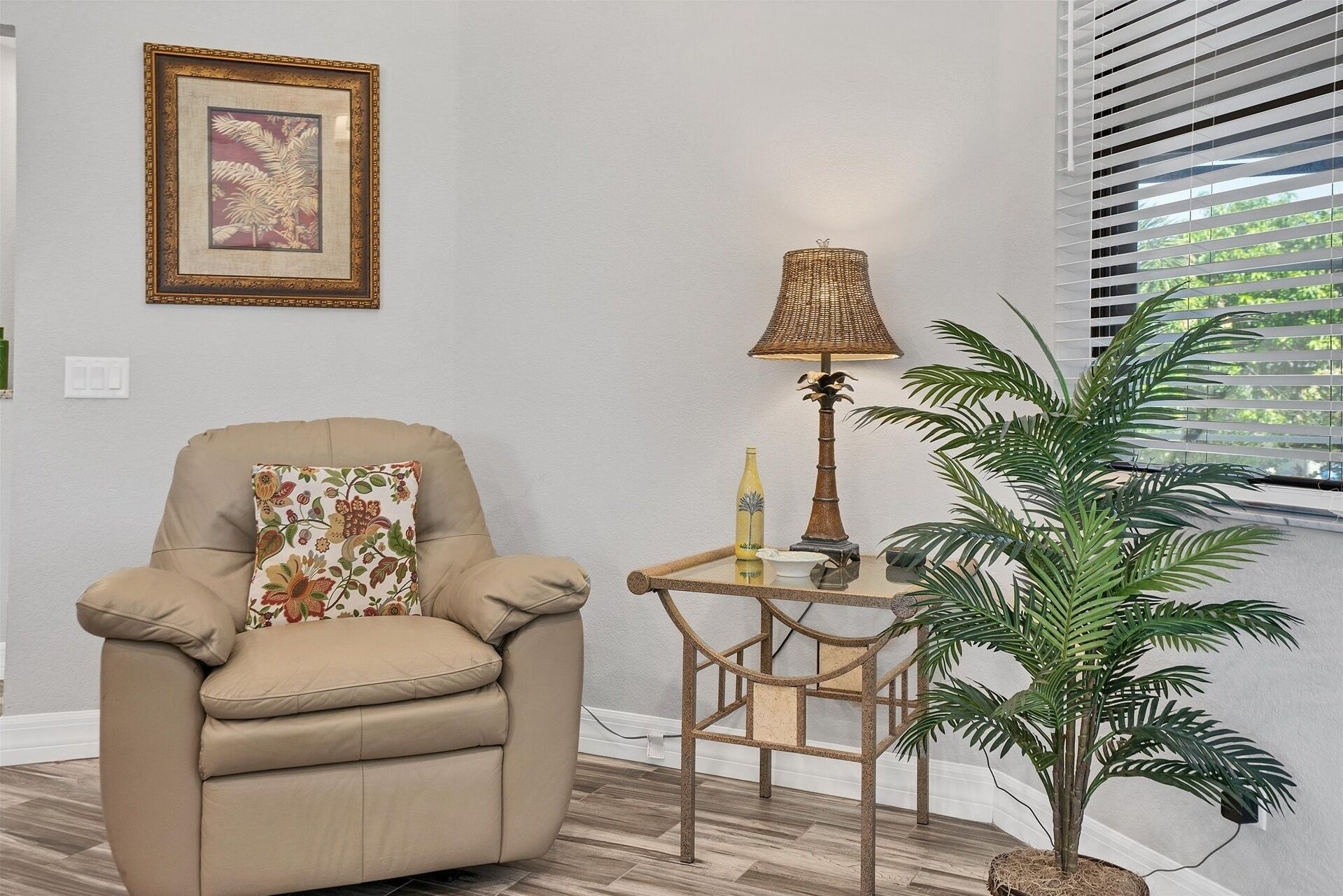Un fauteuil beige agrémenté d’un coussin à motifs floraux, une petite table avec une lampe et des objets décoratifs, et une plante en pot près d’une fenêtre munie de stores.