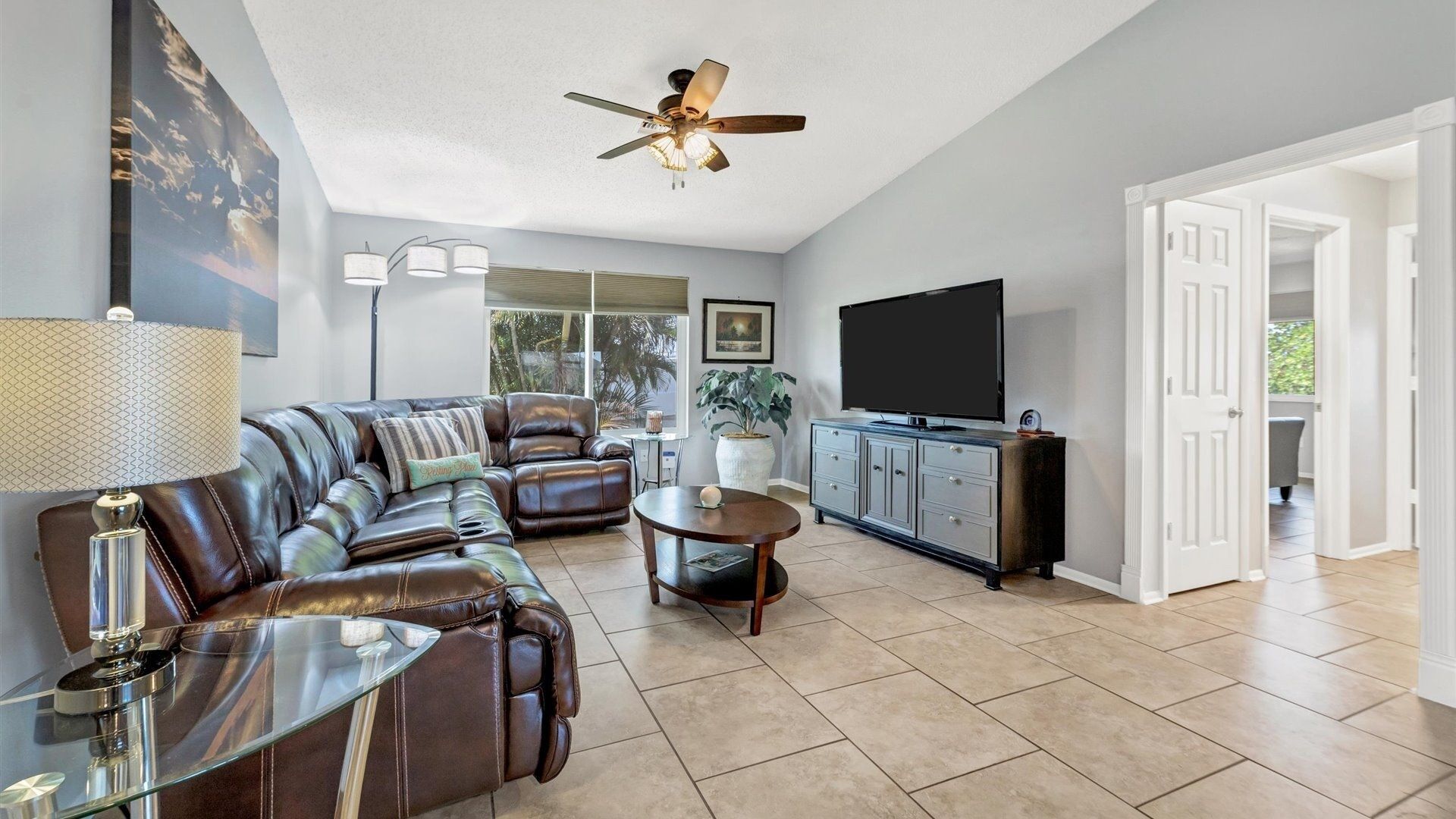A spacious living room with a leather sofa, a glass coffee table, a TV, and a ceiling fan.