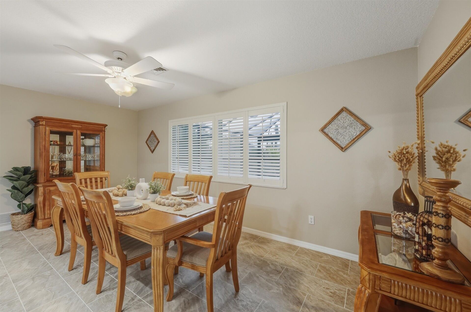 A dining room with a wooden table set for four, surrounded by chairs. A ceiling fan is visible above, and there are decorative wall pieces and a mirror on the wall.