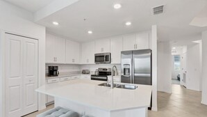 A modern kitchen with white cabinetry, a stainless steel refrigerator, a microwave, and a coffee maker. There are two bar stools at the island. - Heritage Landing Golf And Country Club Vacation Condo 3 Bedroom Condo (Punta Gorda)