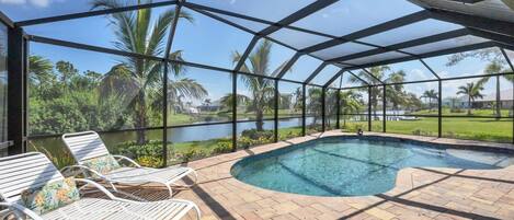 A covered outdoor swimming pool area with lounge chairs and a view of a lake and palm trees.