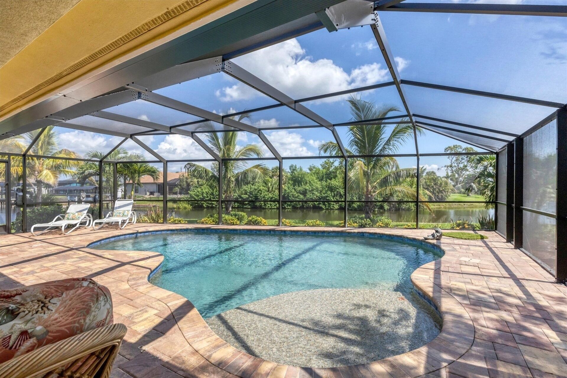 A covered swimming pool with a view of a lake and palm trees, surrounded by lounge chairs and a patio area.