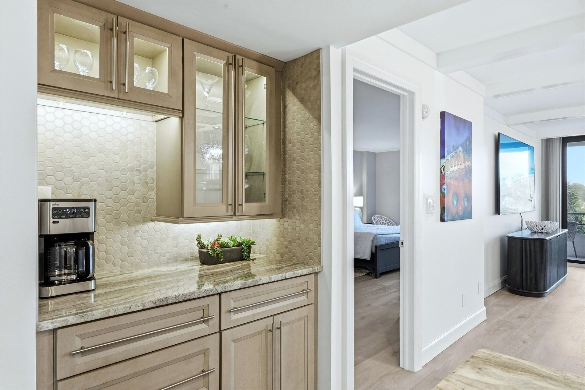 A modern kitchen with a coffee maker, a plant on the counter, and a view into the adjacent bedroom.