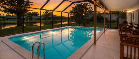 A swimming pool with a glass enclosure, surrounded by outdoor furniture and a view of a sunset.
