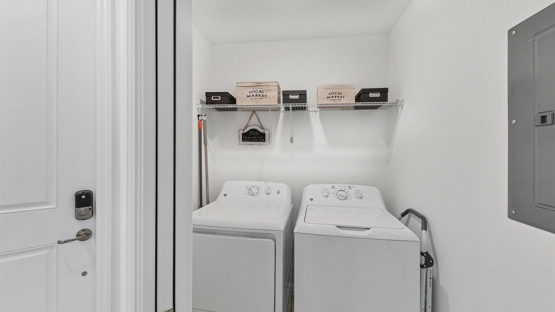 A laundry room with two white washing machines, a shelf with 'LOCAL MARKET' boxes, and a sign requesting guests to clean up after use.