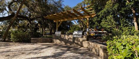 An outdoor barbecue area with a pergola, two grills, and a gravel ground.