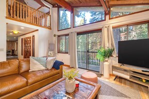 A spacious living room with a brown leather sofa, a wooden coffee table, and a large flat-screen TV. There are multiple windows providing natural light and views of the outdoors. - Aarovan- Pet Friendly, game room (Arnold)