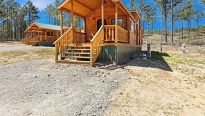 Una cabaña de madera con un porche y escaleras, rodeada de árboles y un cielo despejado.