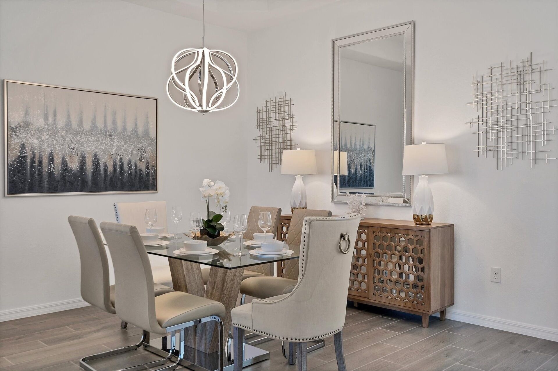A dining area with a glass table, four chairs, a chandelier, a large mirror, and a decorative cabinet.
