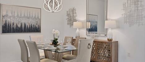 Un comedor con mesa de vidrio, cuatro sillas, una araña, un espejo grande y un aparador decorativo.