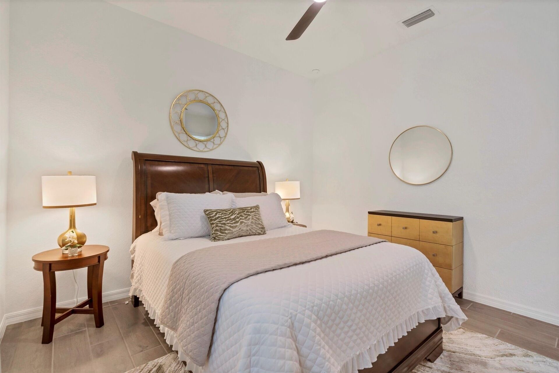 A bedroom with a bed, two bedside tables with lamps, a wooden dresser, and two wall mirrors.