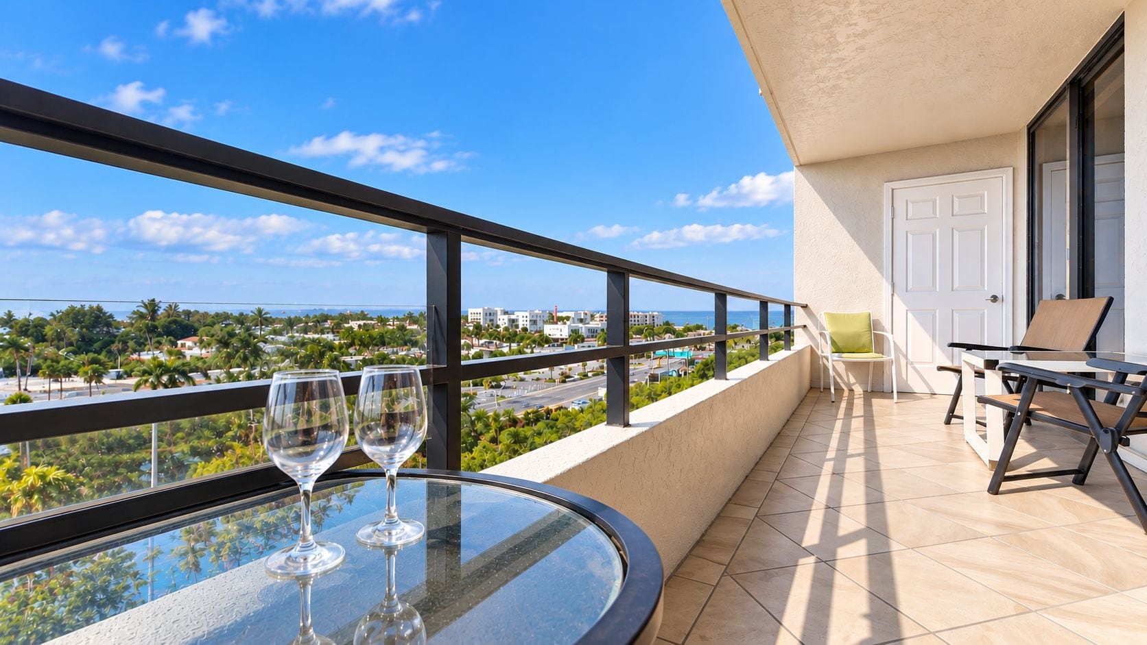 A balcony with a glass table, two wine glasses, a chair, and a view of a coastal area.