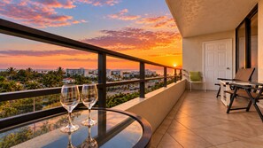 Un balcón con una mesa de vidrio, dos copas de vino, una silla y vistas a un paisaje urbano al atardecer.