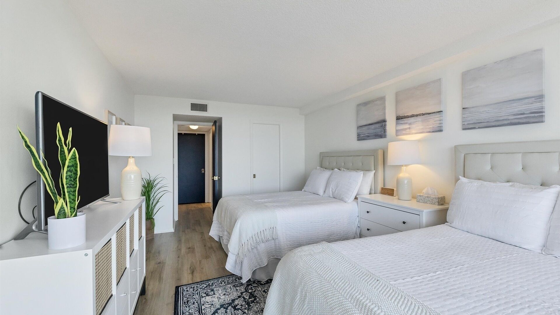 A hotel room with two single beds, a desk with a lamp, a potted plant, and artwork on the walls.
