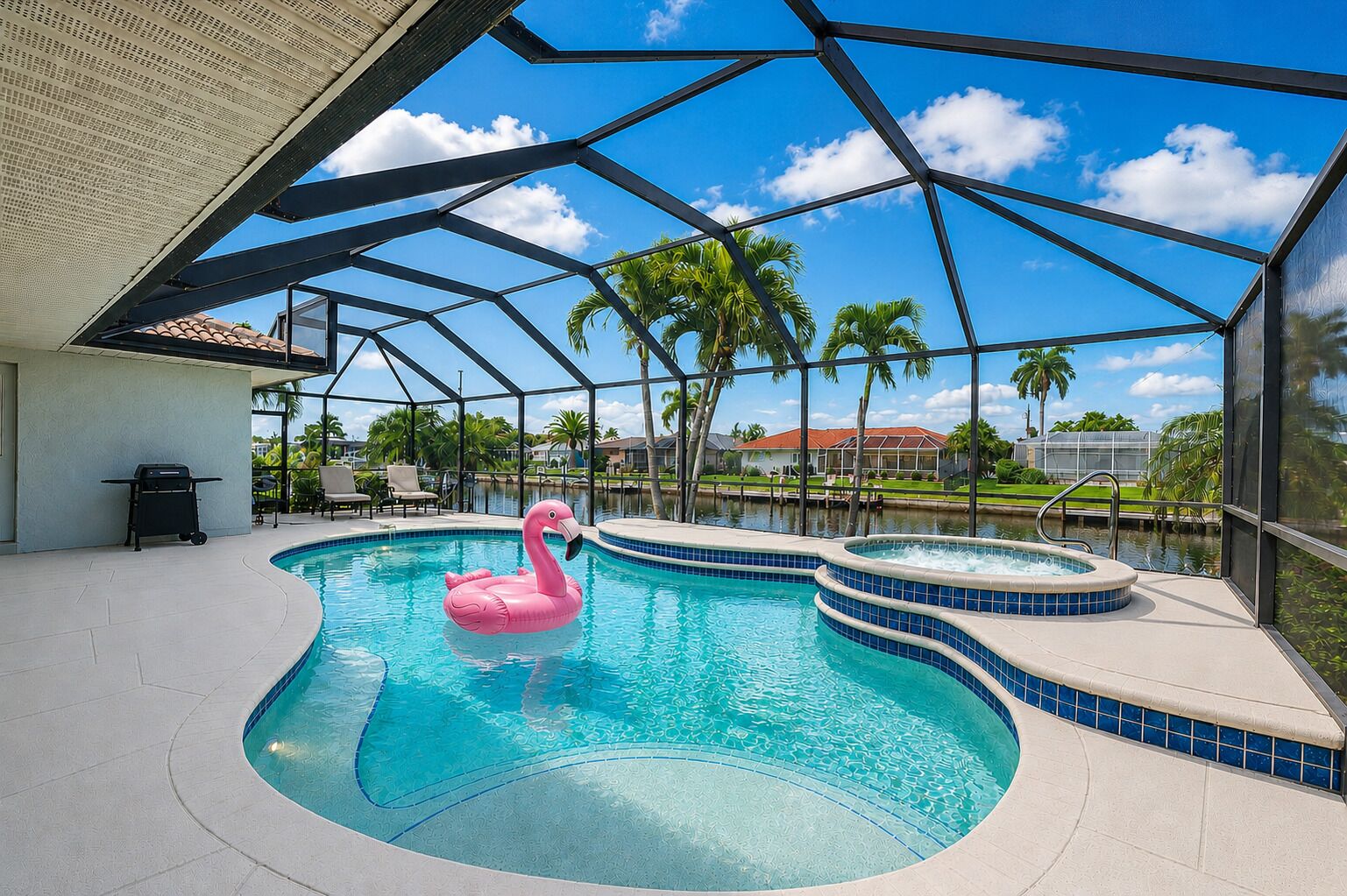 A swimming pool with a pink flamingo float, a hot tub, and a covered patio area with a view of palm trees and houses.