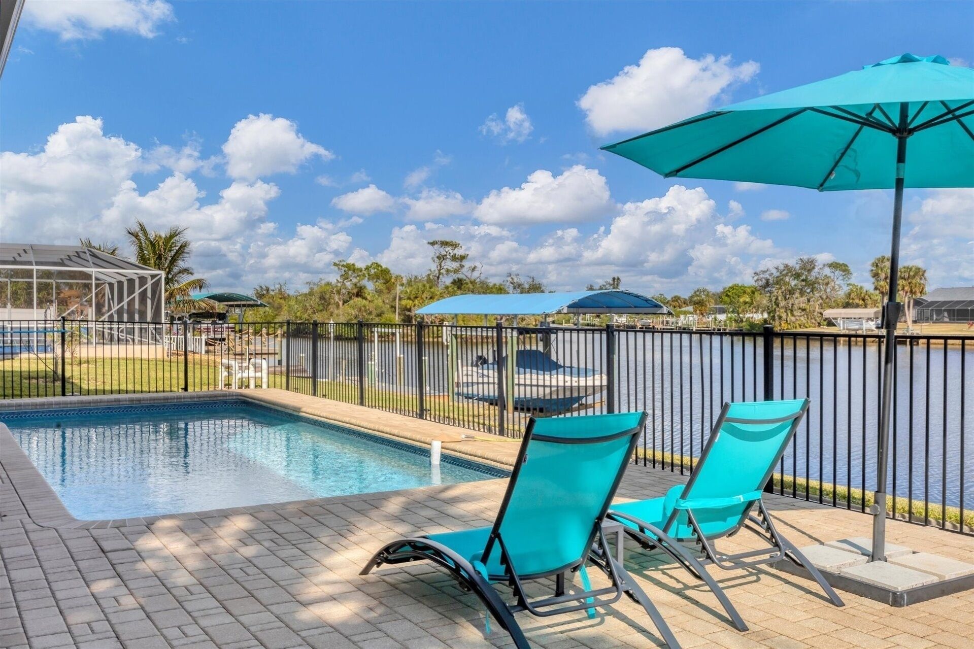 A swimming pool with two turquoise lounge chairs and a matching umbrella, surrounded by a fence and palm trees.