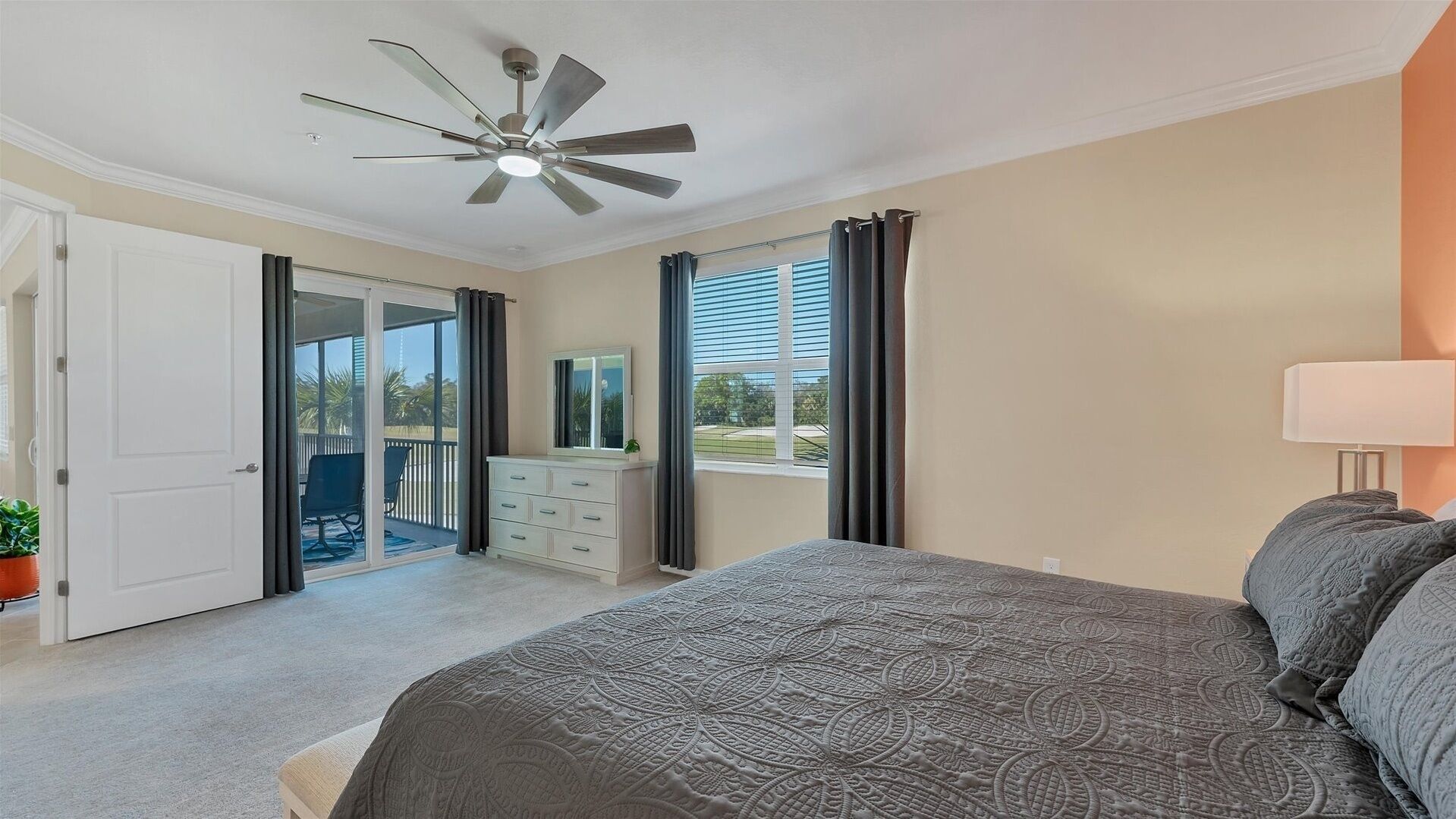 A bedroom with a bed, a ceiling fan, a dresser, a window with curtains, and a view of the outdoors.