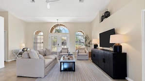 A spacious living room with a large sofa set, a coffee table, and a TV on a black cabinet. There are two windows with arched tops, a ceiling fan, and a door with glass panels. - Punta Gorda Isles 3 Bedroom Pool+Hot Tub Home 2 Home (Punta Gorda)