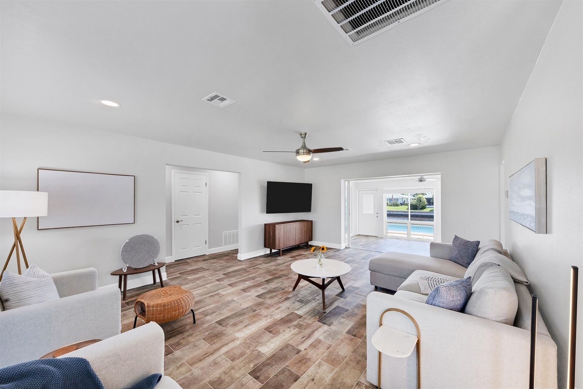 A spacious living room with a large sofa, a coffee table, and a TV mounted on the wall. There is a ceiling fan and a door leading to a balcony.