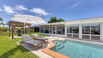 Uma área moderna de piscina exterior com duas espreguiçadeiras, uma pérgula com guarda-sol e um alpendre coberto com churrasqueira.