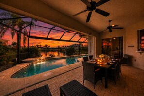 Ein Essbereich am Pool mit Blick auf einen Sonnenuntergang über dem Wasser, einem Deckenventilator und Sitzgelegenheiten im Freien.