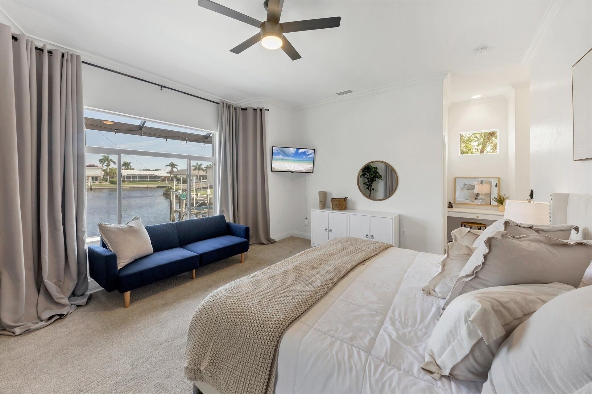 A bedroom with a bed, a blue sofa, a TV, a ceiling fan, a window with curtains, and a view of a body of water.