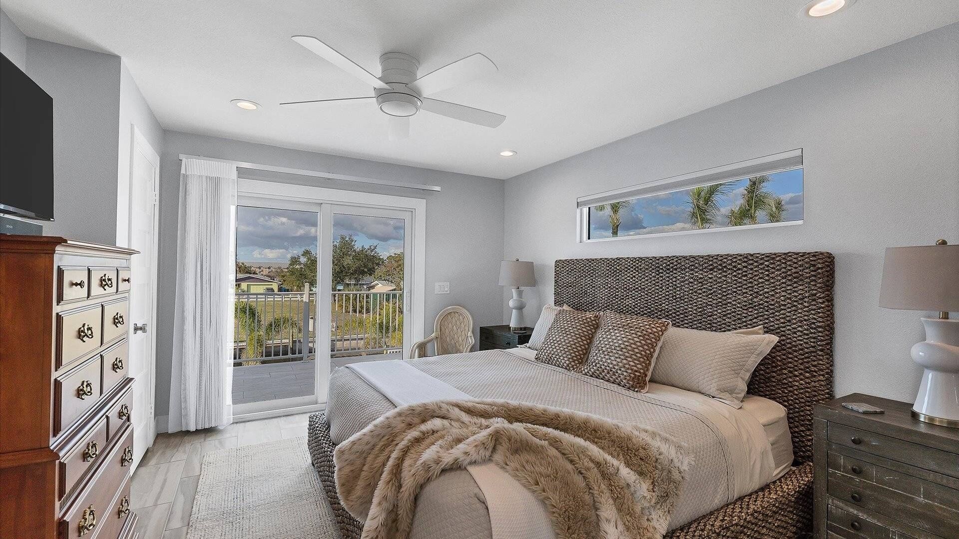 A bedroom with a bed, a ceiling fan, a nightstand, a dresser, and a window with a view of palm trees.