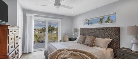 A bedroom with a bed, a ceiling fan, a nightstand, a dresser, and a window with a view of palm trees.