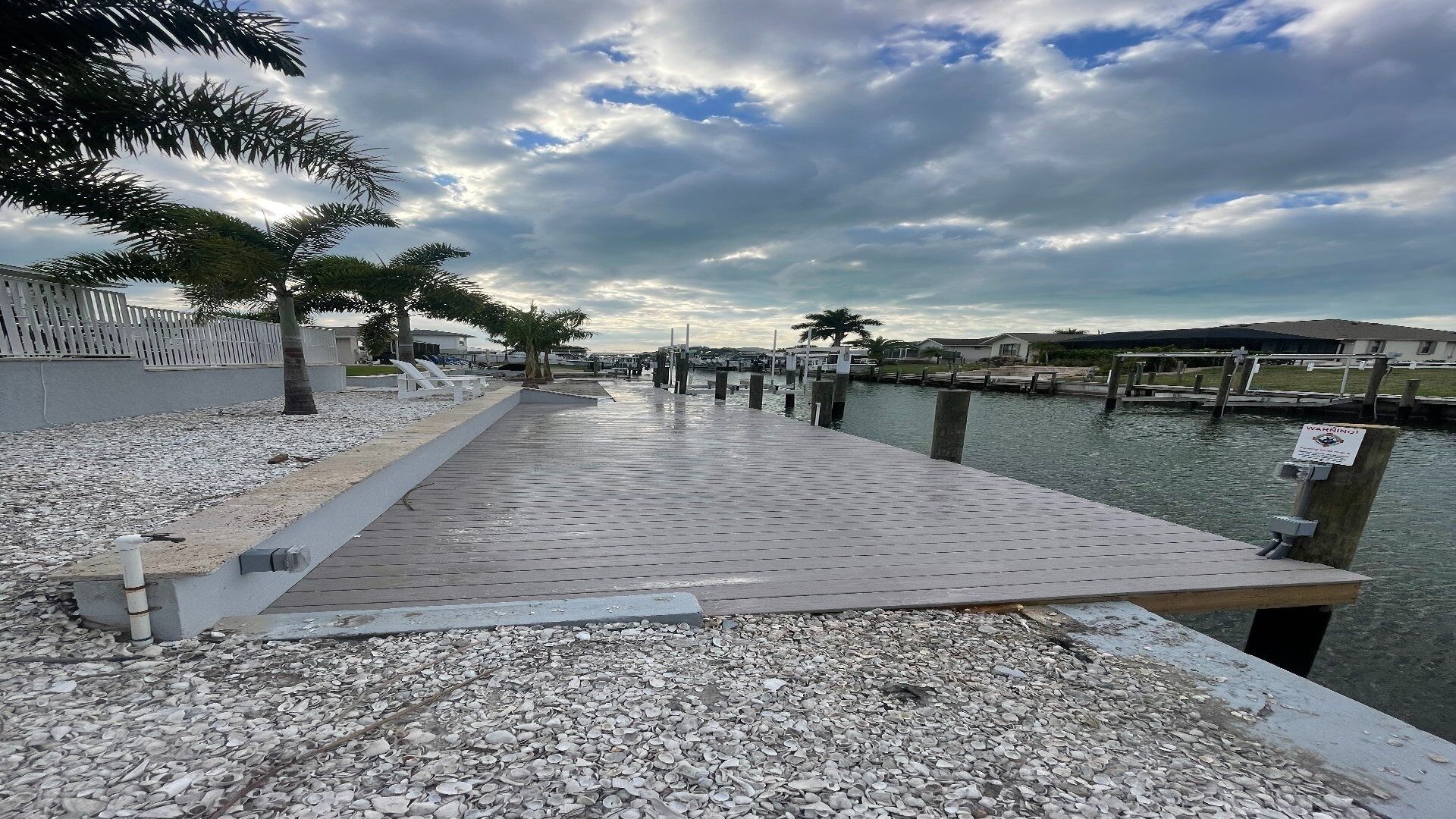 Un paseo marítimo con muelle de madera, piedras, palmeras y una masa de agua tranquila.
