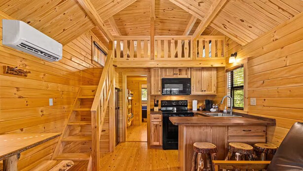 Ein Blockhaus-Interieur mit Holzausstattung, einer Küche mit Mikrowelle, Herd und Spüle, einer Holztreppe und einer Bar mit Hockern.