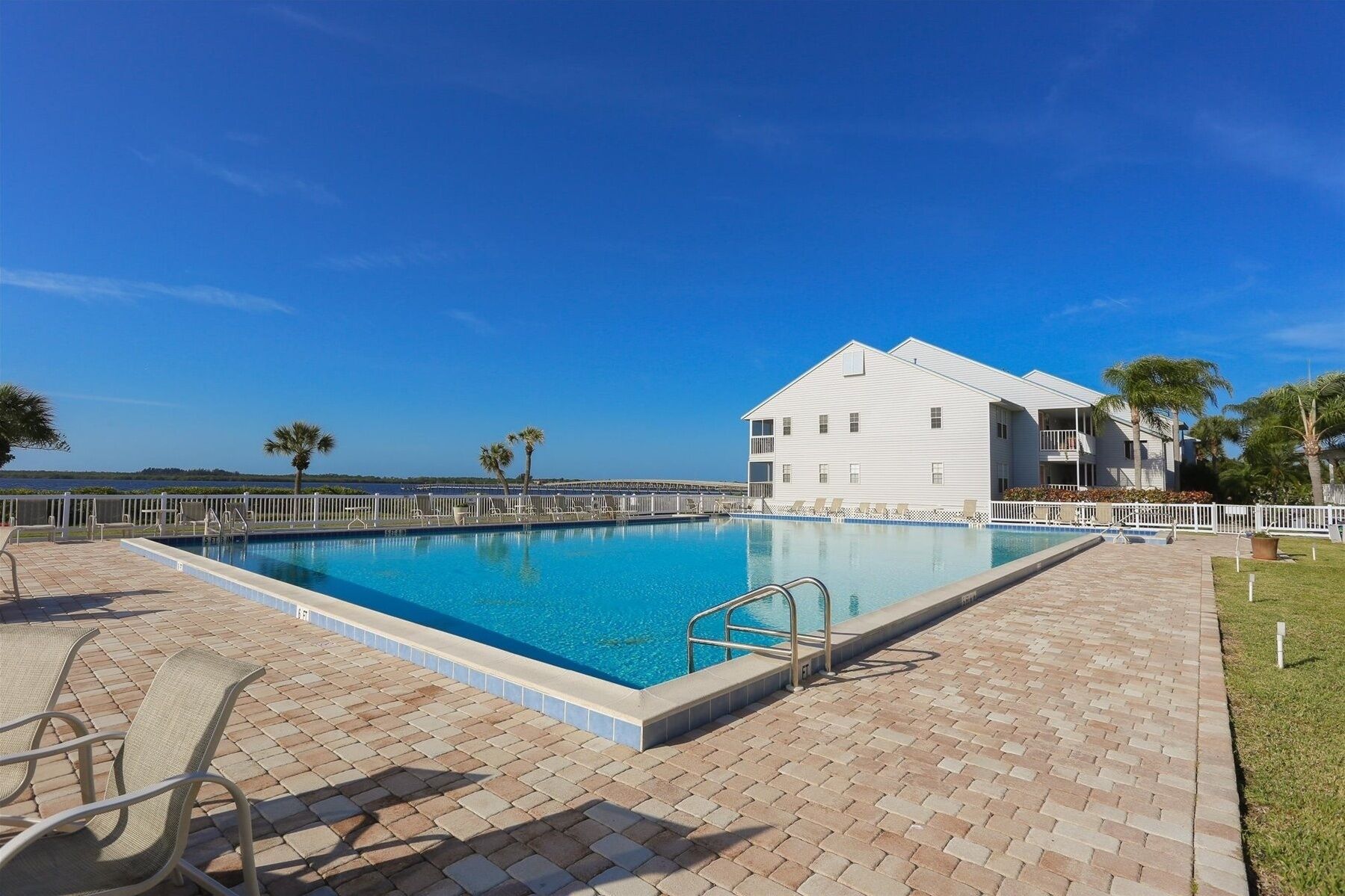 A swimming pool with a clear blue sky, a white building with balconies, and palm trees in the background.