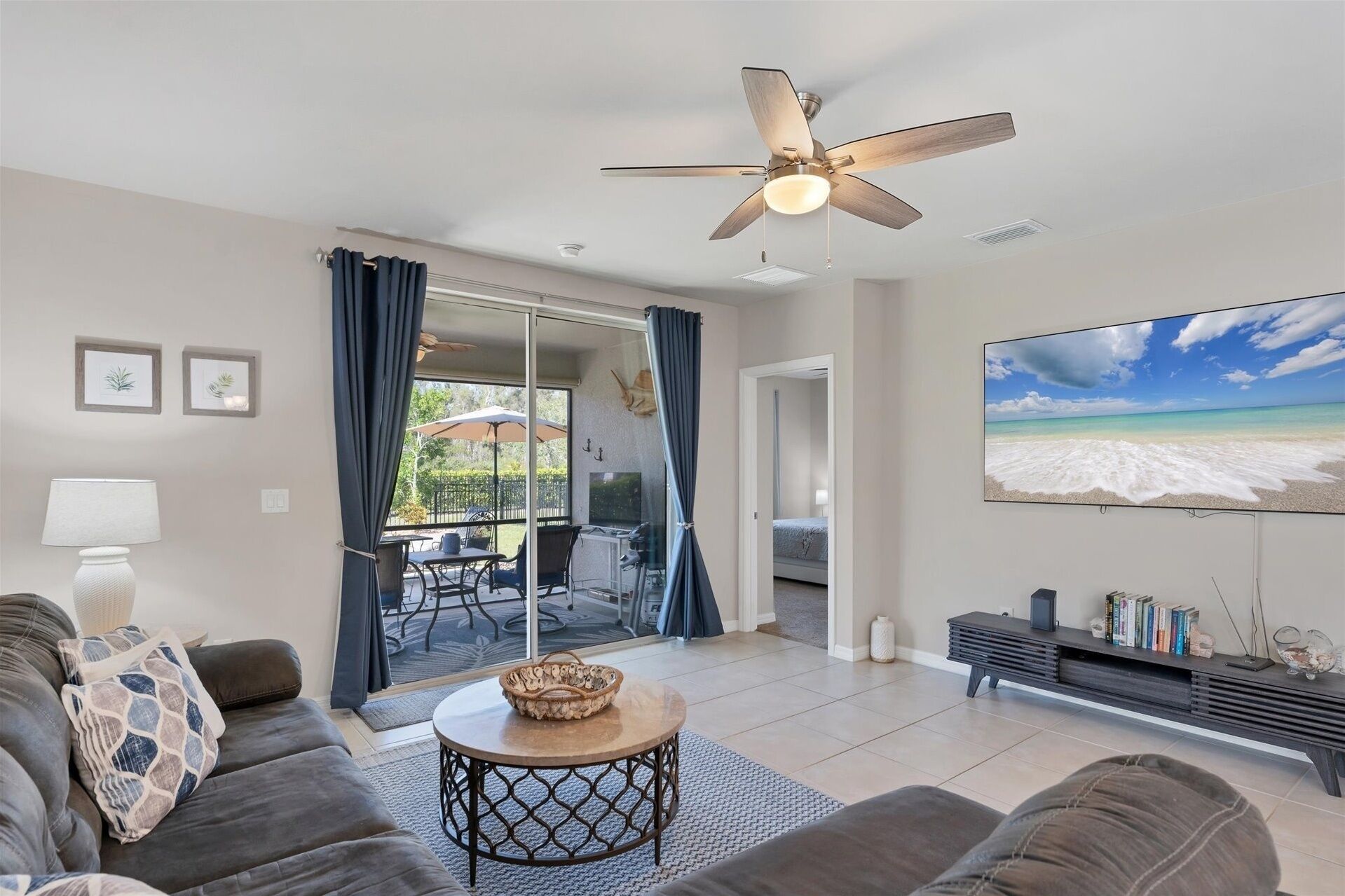 A modern living room with a ceiling fan, a large TV displaying a beach scene, a sofa, a coffee table, and a dining area with a table and chairs.