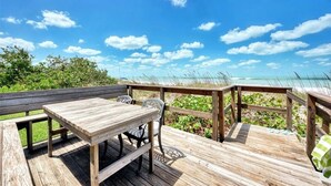 Terraza de madera con mesa y sillas, con vista a una playa y cielos despejados.
