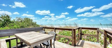 A wooden deck with a table and chairs, overlooking a beach with clear skies.