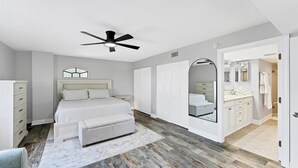 Un dormitorio con cama, ventilador de techo, mesita de noche, cómoda y baño visible a través de una puerta abierta.
