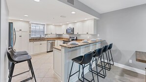 Una cocina moderna con armarios blancos, encimera de granito y microondas integrado. Hay dos taburetes de bar y una isla grande con un fregadero.