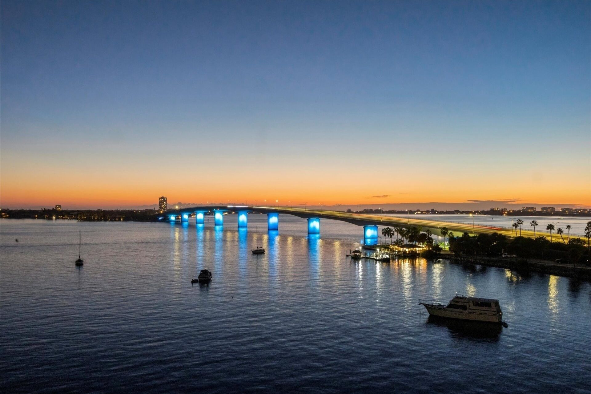 Un pont illuminé de lumières bleues enjambant une étendue d’eau, avec un bateau au premier plan et une vue sur les gratte-ciel d’une ville en arrière-plan.