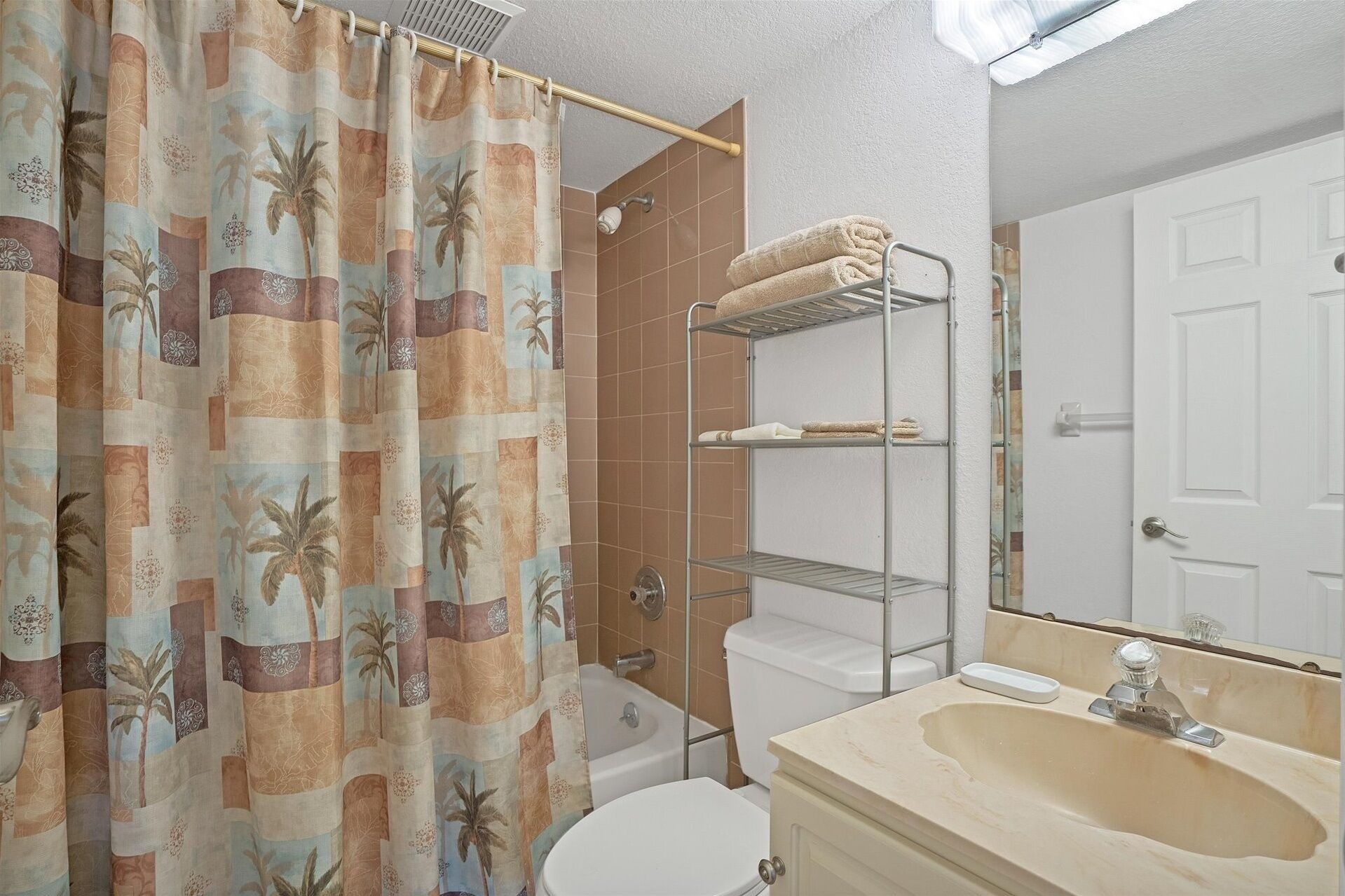 Une salle de bain avec un rideau de douche orné d’un motif tropical, des toilettes, un lavabo avec un miroir et un porte-serviettes avec des serviettes.