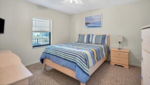 Een slaapkamer met een bed, nachtkastje, dressoir, plafondventilator, raam met jaloezieën en een schilderij aan de muur.