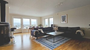 A living room with a black wood-burning stove, a black sofa set, a coffee table, and a large window with a view of the outdoors. - Modern villa with sea view in Svanesund | SE09101 (Svanesund)