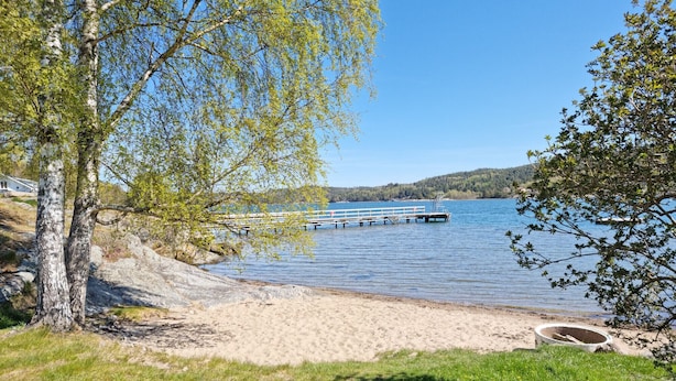 A lakeside scene with a wooden dock extending into the water, surrounded by trees and a sandy beach. - Modern villa with sea view in Svanesund | SE09101 (Svanesund)