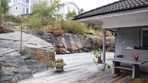 A modern house with a wooden deck, a stone wall, and a garden with potted plants. - Modern villa with sea view in Svanesund | SE09101 (Svanesund)