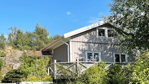 Une maison en bois rustique, dotée d’un porche, entourée de verdure et surplombée d’un ciel bleu azur.