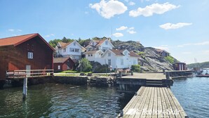 Un muelle de madera que lleva a un embarcadero con una hilera de casas tradicionales, una ladera rocosa y un cielo azul despejado.