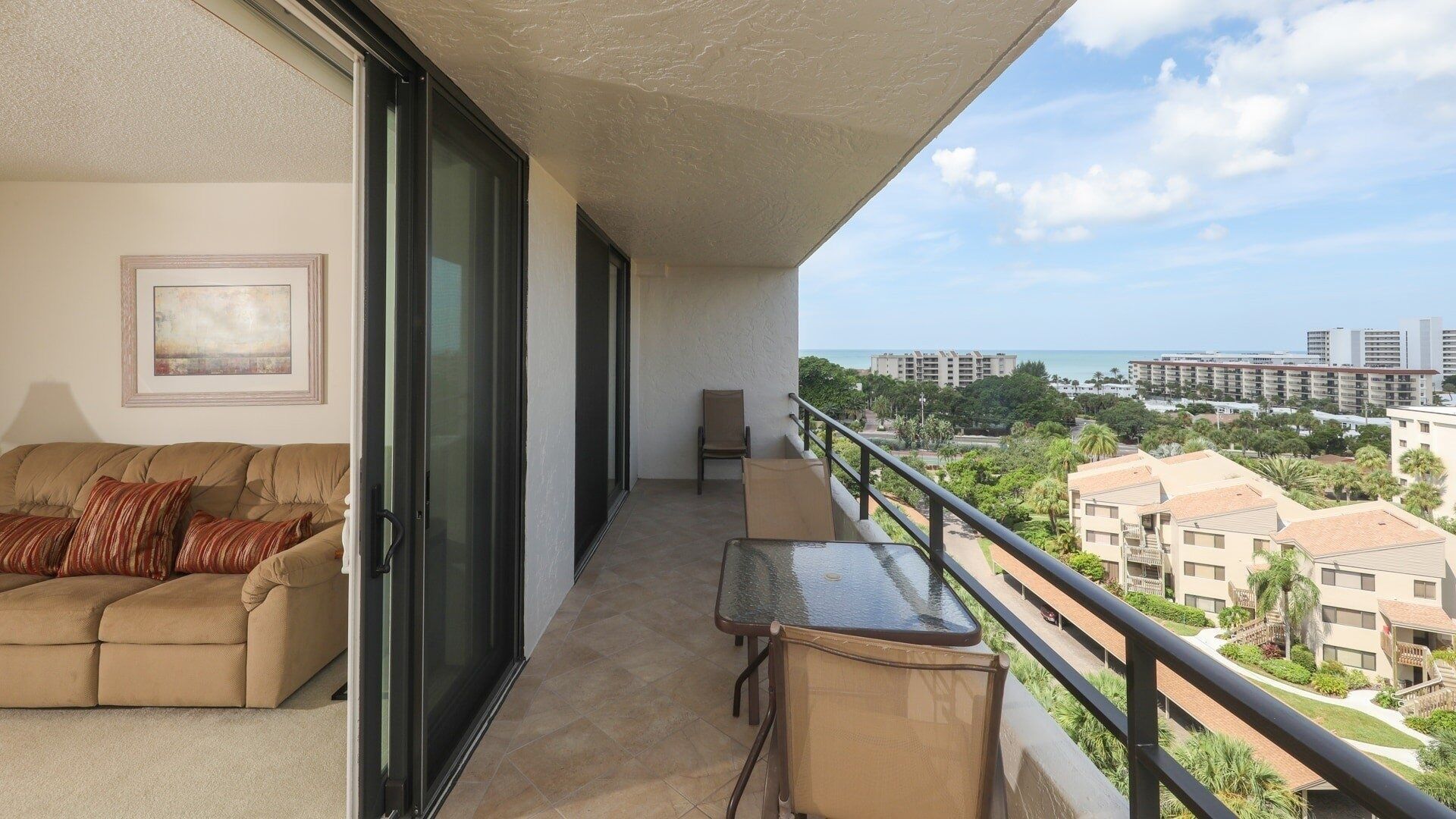 A balcony with a sofa and a table, a chair, and a view of a residential area and the sea.